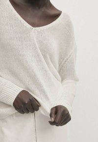 Biały, luźno dziergany sweter z szerokim dekoltem w serek i prążkowanymi mankietami. Lekka, teksturowana tkanina. Model trzyma dół swetra, prezentując swobodny krój.