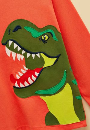 Helepunane kangas suure rohelise dinosauruse aplikatsiooniga, kus on avatud suu, valged hambad, punane keel ja kollakasrohelised detailid.