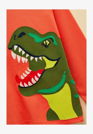 Kirkkaan oranssi kangas, jossa suuri vihreä dinosaurusapplikaatio avoin suu, valkoiset hampaat, punainen kieli ja kelta-vihreät korostukset.