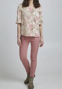 Bluse mit floralem Print in Beige, Pink und Grün; mit kurzen Ärmeln und V-Ausschnitt. Kombiniert mit enganliegenden pinken Hosen und grünen Stiefeletten.