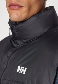 Black insulated vest with a high collar, front zipper, and a white logo on the left chest. Smooth texture and lightweight design.