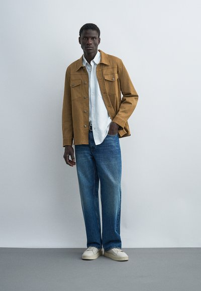 Άνδρας με καφέ μπουφάν με κουμπιά πάνω από λευκό πουκάμισο, μπλε τζιν και λευκά αθλητικά παπούτσια, στέκεται μπροστά σε έναν απλό φωτεινό τοίχο.