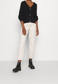 Cardigan nero a maglia con scollatura a V e bottoni, abbinato a pantaloni effetto avorio arrotolati e stivaletti neri. Design semplice e testurizzato.