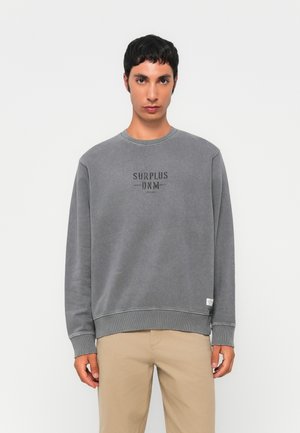 Young man wearing a faded grey sweatshirt with "SURPLUS DNM" text and beige pants, standing against a plain white background.