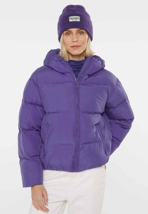 Frau trägt eine violette Steppjacke mit Reißverschluss, passende gestrickte Mütze mit Aufschrift "SENSES THE LABEL," und weiße Hose vor einfachem Hintergrund.