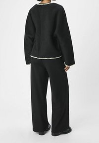 Person som bär svarta vida byxor och en svart texturerad jacka med vit fläktkant på kragen, ärmarna och nederkanten, står mot en vit bakgrund.