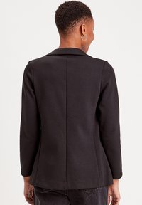 Blazer noir au design ajusté, avec un col à revers, des manches longues et une texture lisse. Le dos présente un détail de couture central.