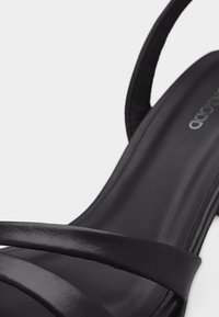 Μαύρο δερμάτινο σανδάλι με λεπτούς σταυρωτούς ιμάντες και χαμηλό τακούνι, που αναδεικνύει λεία υφή και μινιμαλιστικές σχεδιαστικές λεπτομέρειες.