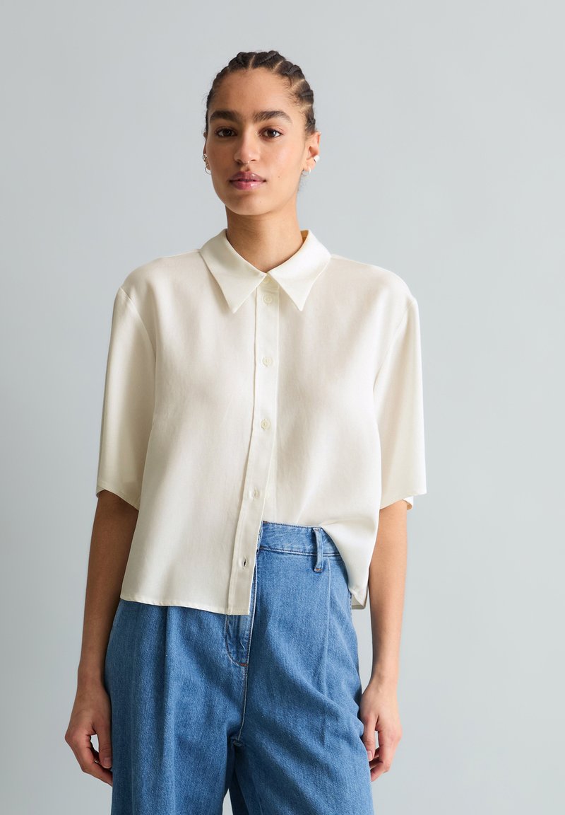 Γυναίκα με πλεξούδες φοράει ένα λευκό πουκάμισο με κοντά μανίκια και κουμπιά, κεντημένο σε ψηλόμεσο μπλε τζιν, μπροστά σε απλό γκρίζο φόντο.