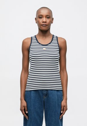 Person wearing a sleeveless navy and white striped top with a small tiger emblem, paired with high-waisted loose blue jeans, standing facing forward.