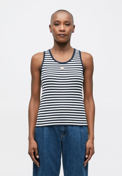 Eine Person trägt ein ärmelloses, marineblau und gestreiftes Oberteil mit einem kleinen Tiger-Emblem, kombiniert mit hochgeschnittenen, lockeren blauen Jeans, und steht frontal.