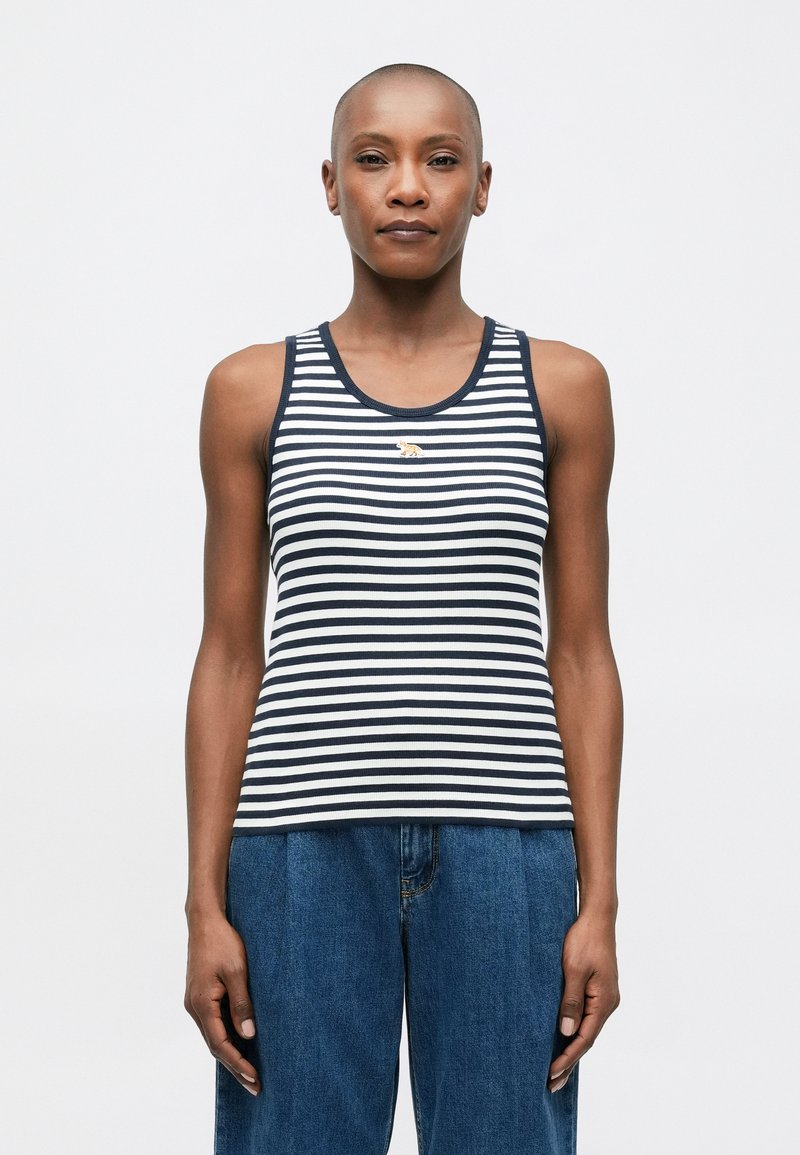 Cilvēks, kurš valkā bezdelņu, tumši zilu un baltu svītrainu kreklu ar mazu tīģera emblēmu, apvienojot to ar augstās jostasvietas brīviem ziliem džinsiem, stāv vērsts uz priekšu.