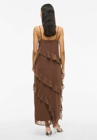 Vrouw in een mouwloze bruine maxi-jurk met diagonale ruches, staande met haar rug naar de camera, met witte hakken aan.