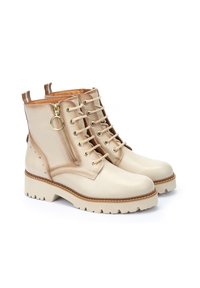 Botas de cuero beige hasta el tobillo con suela gruesa, cordones frontales, cremallera lateral y tachuelas decorativas en los laterales. Textura suave con acentos marrones contrastantes.