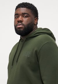 Green hoodie with a drawstring, soft texture, and a subtle reflective logo. Features a relaxed fit and ribbed cuffs.