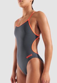 Vrouw in een donkergrijs badpak met oranje details en het "arena" logo, staande tegen een lichtgrijze achtergrond.