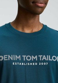 Tirkīza kokvilnas T-krekls ar izteiksmīgu baltu tekstu "DENIM TOM TAILOR" un zem tā rakstīts "ESTABLISHED 2007" mazākā fontā. Apaļā kakla izgriezums.