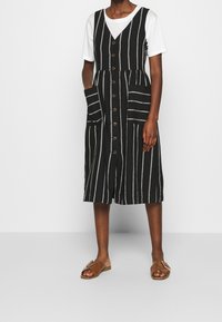 Robe rayée noir et blanc avec un devant à boutons, deux grandes poches et un corsage ajusté. Portée sur un t-shirt blanc, elle présente un ourlet longueur genou.