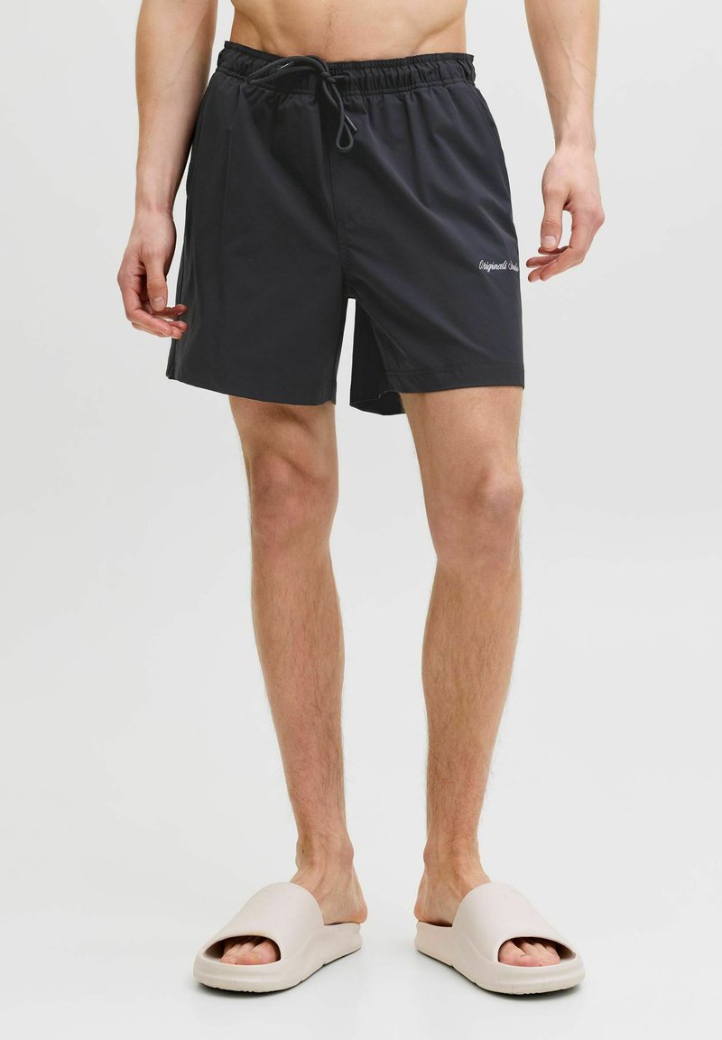 Homme portant un short de bain noir à cordon et des claquettes beige à bout ouvert, debout devant un fond clair uni.