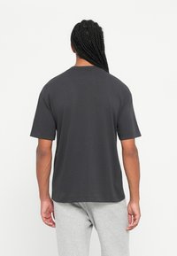 Άνδρας με μακριά πλεξίμ�ατα φορώντας ένα απλό σκούρο γκρι κοντομάνικο μπλουζάκι και ανοιχτό γκρι παντελόνι, στέκεται στραμμένος προς τα πίσω.