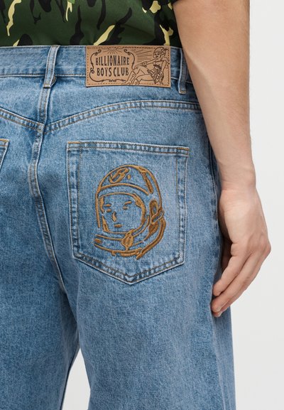 Farmernadrág barna hímzett űrhajós sisakkal a hátsó zsebén és bőr címkével, amelyen a Billionaire Boys Club felirat olvasható.