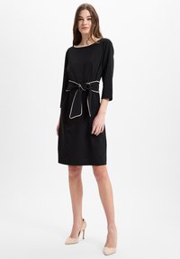 Schwarzes Kleid mit Dreiviertelärmeln, einer figurbetonten Silhouette und einer in einer kontrastierenden hellen Farbe vorne gebundenen Taille.
