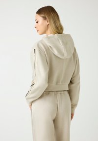 Beżowa krótka bluza z kapturem, z czarnymi paskami po bokach i fakturą ryżową. Osoba jest pokazana z tyłu, w luźnych spodniach.