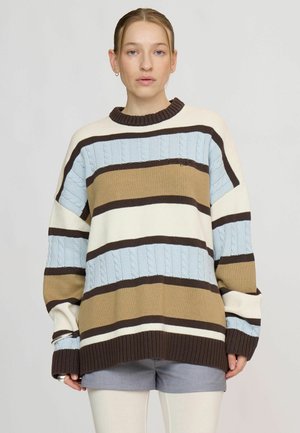 Jeune personne portant un pull ample rayé bleu, marron, crème et marron foncé, associé à un short gris et des leggings blancs.