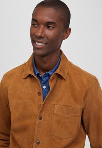 Chaqueta de ante marrón con cierre de botones, bolsillo en el pecho y una textura suave, superpuesta sobre una camisa de mezclilla azul con cuello.