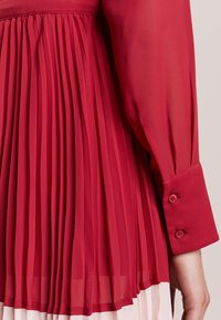 Blusa roja con plisados y tela translúcida, de mangas largas y puños abotonados; presenta textura vertical y un diseño suave y fluido.
