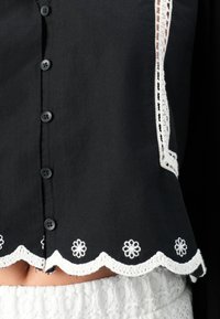Blouse en coton noir avec un ourlet festonné orné de dentelle blanche au crochet et d'accents de fleurs brodées, agrémentée de boutons noirs.