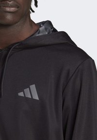 Svart hoodie med en slät yta, prydd med en grå Adidas-logotyp på bröstet och ett mönstrat innerhuvud. Justerbar dragsnodd.