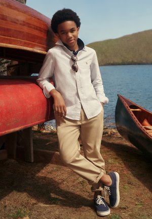 Jeune personne appuyée contre des canoës empilés au bord d'un lac, portant une chemise rayée, un pantalon beige et des baskets bleu marine avec des lunettes de soleil accrochées à la chemise.
