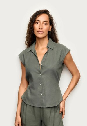 Donna con capelli ricci che indossa una camicia senza maniche verde oliva con bottoni e pantaloni larghi abbinati, in piedi con una mano sui fianchi.
