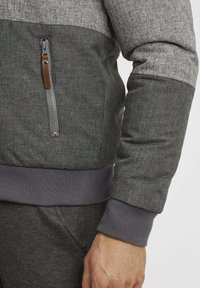 Gray quilted jacket with a textured finish, featuring a zippered pocket and ribbed cuffs. Highlighted by a contrasting darker lower section.
