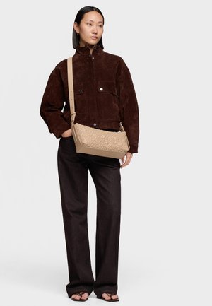 Kobieta o prostych czarnych włosach, ubrana w ciemnobrązową zamszową kurtkę i spodnie, nosząca beżową pikowaną torebkę na ramię, stojąca z jedną ręką w kieszeni.