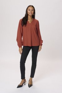 Roestkleurige blouse met een v-hals, een gestructureerd patroon en lange mouwen; gecombineerd met zwarte slim-fit broeken en zwarte hakken.