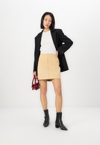 Blazer nero, top bianco e gonna beige. Stivali neri alla caviglia con punte appuntite. Tiene una piccola borsa rossa. Design semplice e moderno.