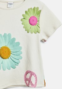 Krátky rukáv krémového trička s zelenou a modrou kvetinovou výšivkou, s ružovým symbolom pokoja a prúžkovanou textúrou. Viditeľná značka.