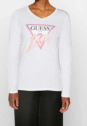 Person trägt ein weißes Langarm-V-Ausschnitt-Shirt mit einem pink-schwarzen "GUESS ICON"-Logo, kombiniert mit einer schwarzen Hose.