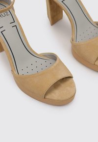 Sandalias de tacón abiertas en la parte delantera de ante beige, con tacón bloque, que presentan una amplia correa frontal y una suela interna lisa con detalles perforados.