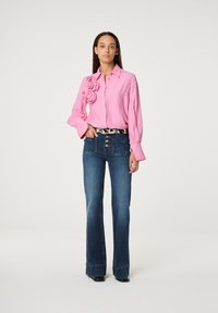 Roze blouse met bloemenaccenten aan de linkerkant, gecombineerd met wijde blauwe denim jeans en een gestreepte zwart-witte riem. Soepele texturen zichtbaar.