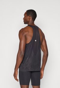 Μαύρο αθλητικό τοπ με διάτρητο σχέδιο και χαμηλή πλάτη, διαθέτοντας μια μικρή ανακλαστική λεπτομέρεια. Συνδυάζεται με μαύρες εφαρμοστές σορτς.