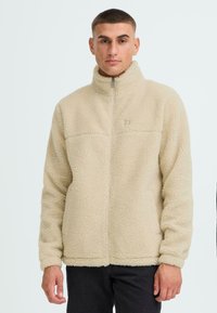 Beige fleece jack met een hoge kraag, ritsluiting en een gestructureerd oppervlak. Heeft een horizontale naad over het borstgebied.