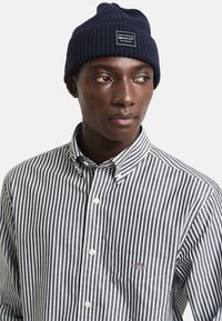 Donkerblauwe geribde gebreide beanie met een klein vierkant label. Draagt een zwart-wit gestreepte overhemd met knopen, met witte knopen.