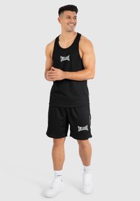 Svart bomullstopp och shorts med "SMILODOX"-text. Toppen har breda ärmhål; shortsen har sidofickor och en ribbad textur. Vita sneakers.