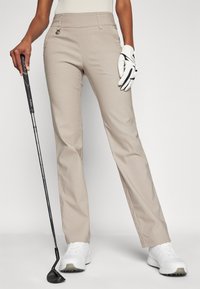 Pantalons de golf beige avec une coupe ajustée, dotés d'une taille et de poches avant. Assortis à des chaussures de golf blanches et un club de golf noir.
