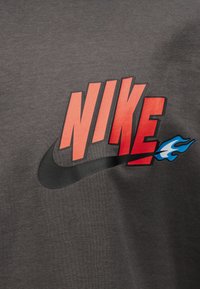 Material gri închis cu text roșu îndrăzneț "NIKE", logo swoosh negru care se extinde cu o mică flacără albastră și albă în partea dreaptă.
