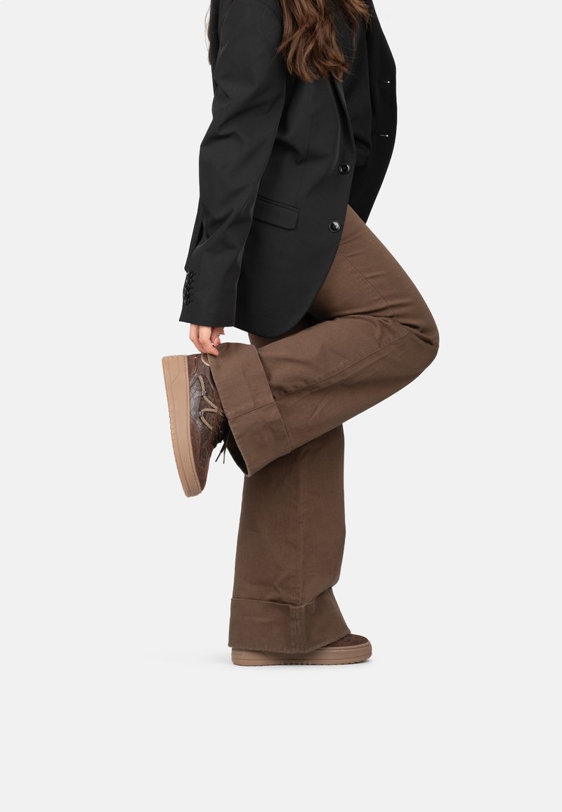Pantaloni a zampa d'elefante in corduroy marrone, con orlo risvoltato, abbinati a sneakers marroni con motivo. Blazer nero con bottoni e dettagli in tessuto.