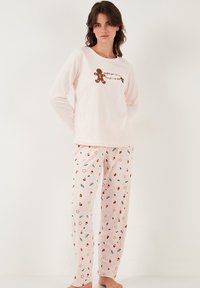 Vaaleanpunainen pyjama, jossa on pitkähihainen paita, jossa on piparkakkutalon kuva ja teksti, yhdistettynä eri juhlallisia kuvioita sisältäviin housuihin.